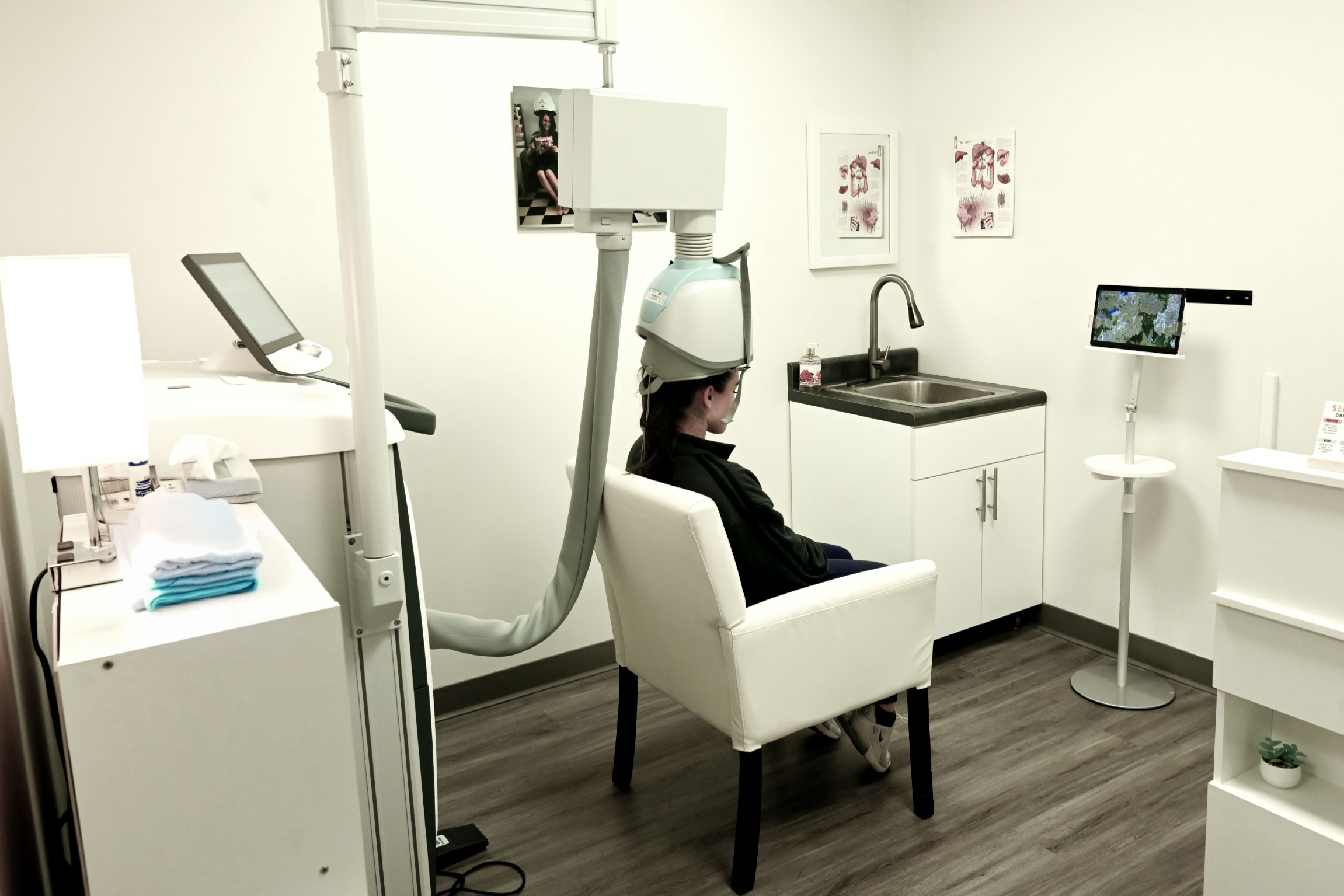 The image shows an individual sitting in a dental chair with their head inside a dental x-ray machine, likely at a dentist's office, where there are other chairs and equipment visible.