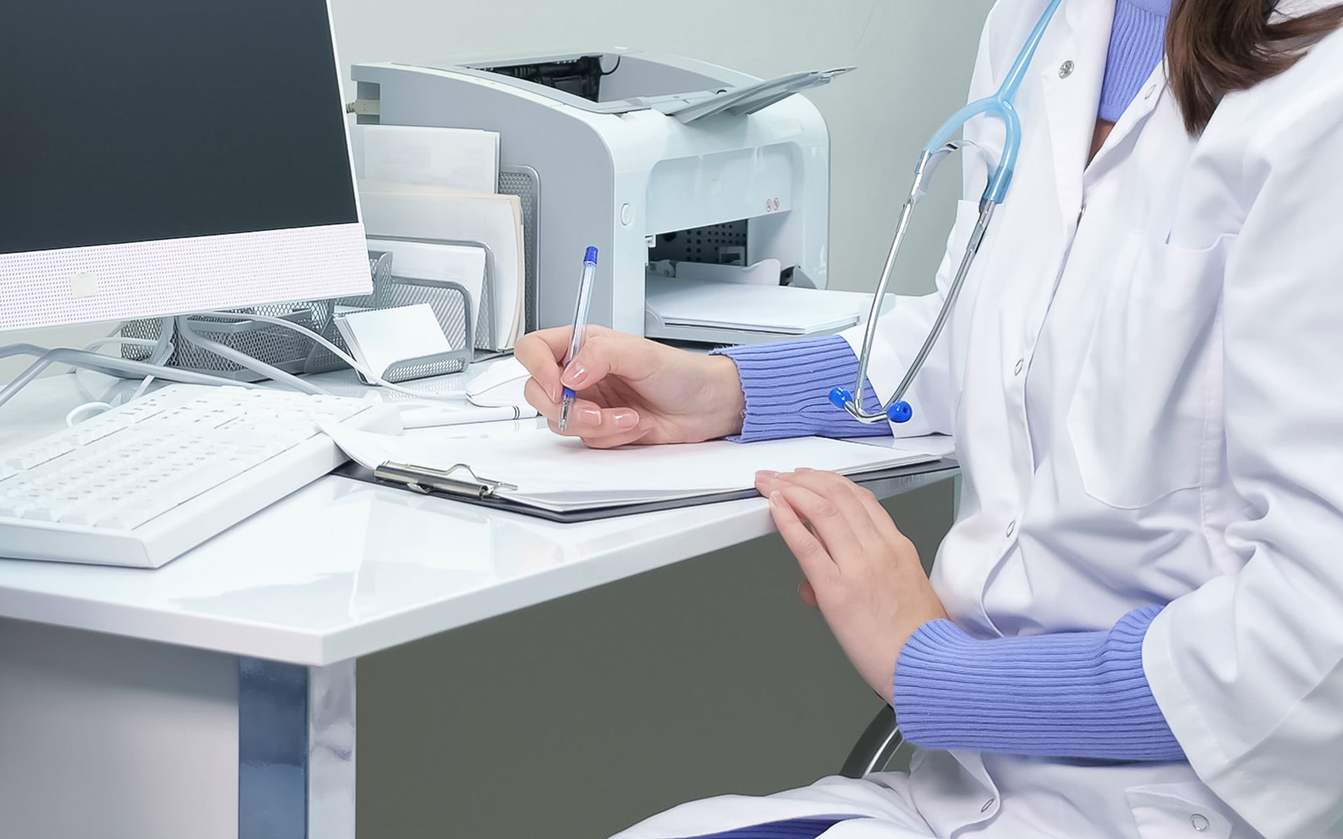 The image shows a healthcare professional seated at a desk with paperwork, wearing a white lab coat and stethoscope, and writing on a clipboard while looking down.