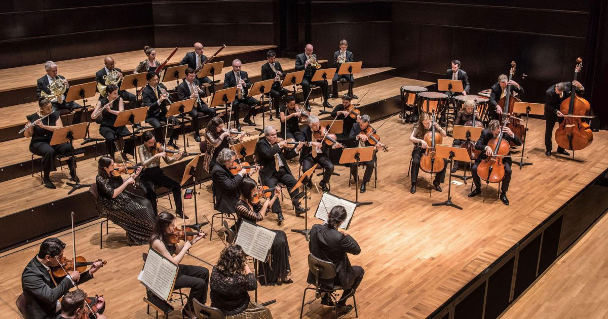 The image shows a symphony orchestra performing on a stage with wood paneling, musicians seated behind music stands, and a conductor leading from the front.