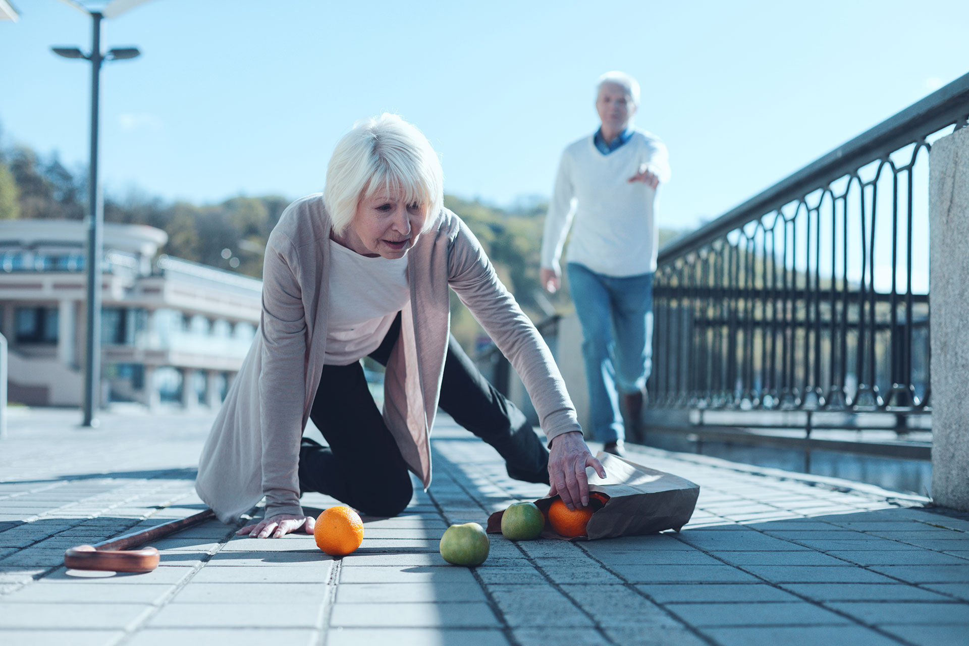 An elderly woman is bending over to pick up fruit on the ground while an older man watches her from behind.
