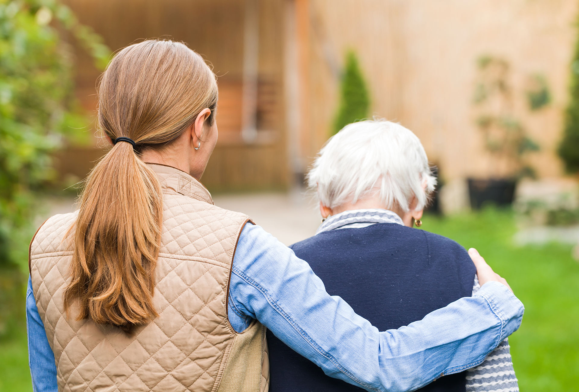 The image shows two individuals standing outdoors with one person appearing to be assisting the other, who may require additional support due to age or mobility issues. They are both dressed casually and seem to be engaged in a supportive interaction.