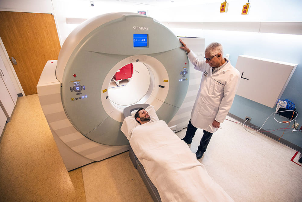 In the image, a man in a white coat stands next to a large, cylindrical medical imaging machine with a red heart on its side, while another person is lying down in front of it. The setting appears to be a hospital room, as indicated by the presence of medical equipment and the professional attire of the individuals.