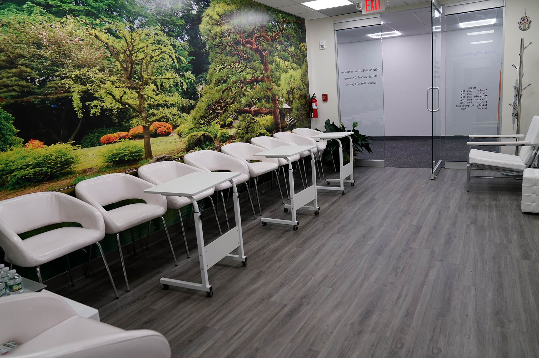 The image shows a modern waiting room with a colorful wall mural, multiple chairs on wheels, and a reception desk with a computer monitor displaying text.