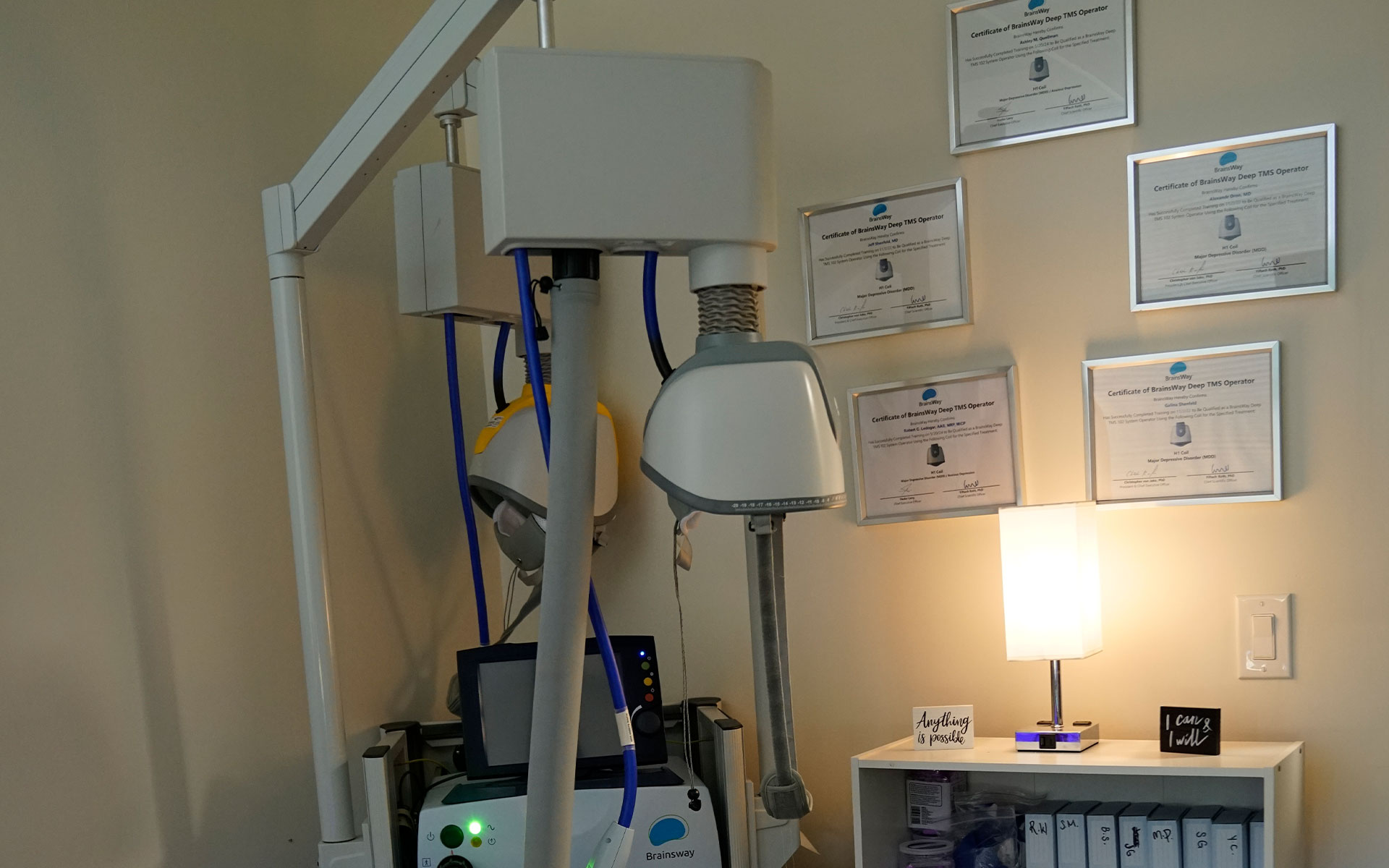 The image shows a dental office setting with various pieces of equipment, including a dental chair, overhead lamp, and what appears to be an X-ray machine, along with certificates on the wall indicating qualifications or accreditation.
