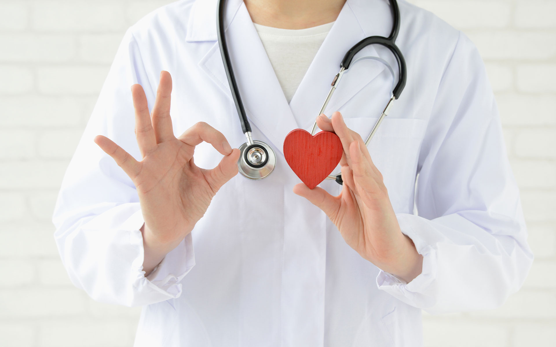 The image features a healthcare professional wearing a white lab coat with a stethoscope around their neck, holding a red heart with both hands while making a peace sign gesture with one hand.
