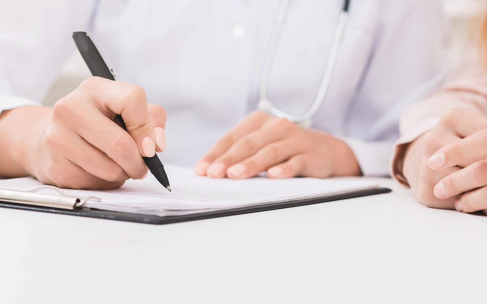 The image shows two individuals, likely healthcare professionals, sitting at a table with a clipboard and pen, focused on reviewing documents, with one person writing on a tablet.