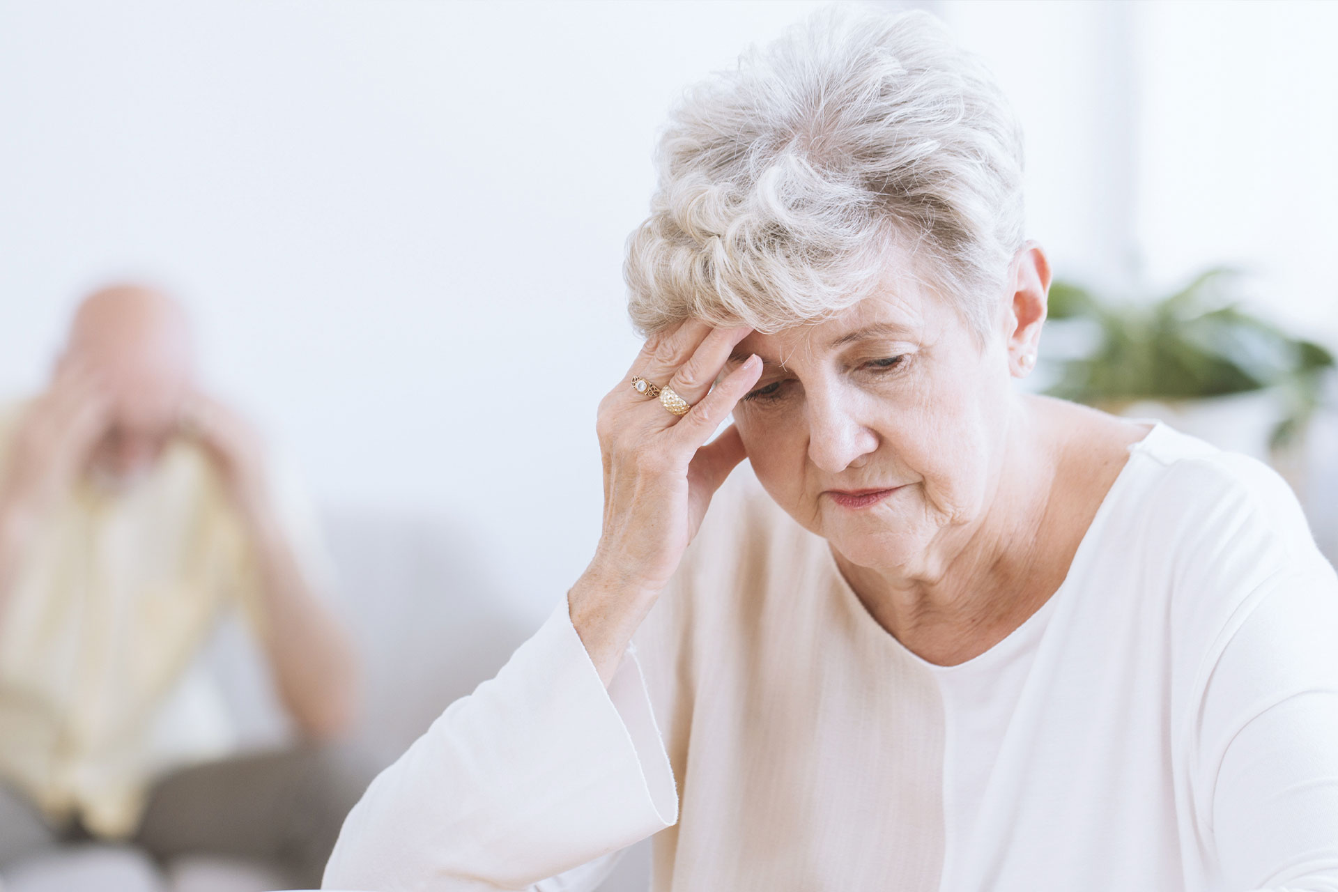 The image shows an elderly woman with her hand on her forehead, appearing distressed or overwhelmed, while another person is visible behind her, seemingly unaware of her condition.