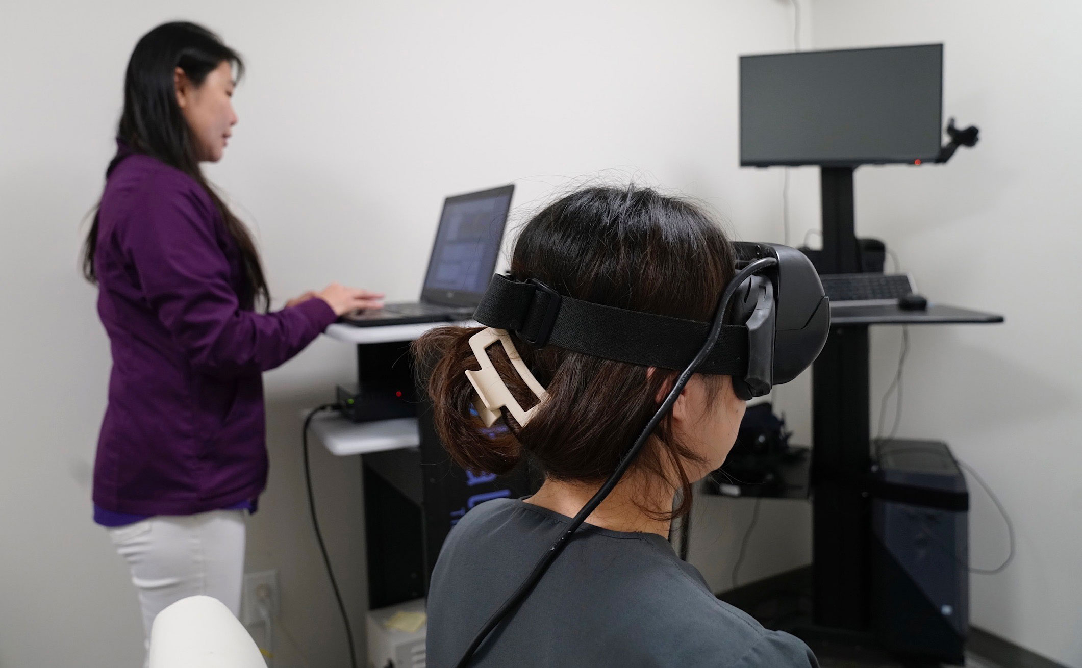 Two individuals seated at a desk with a computer monitor and VR headset, engaged in an activity requiring focus.