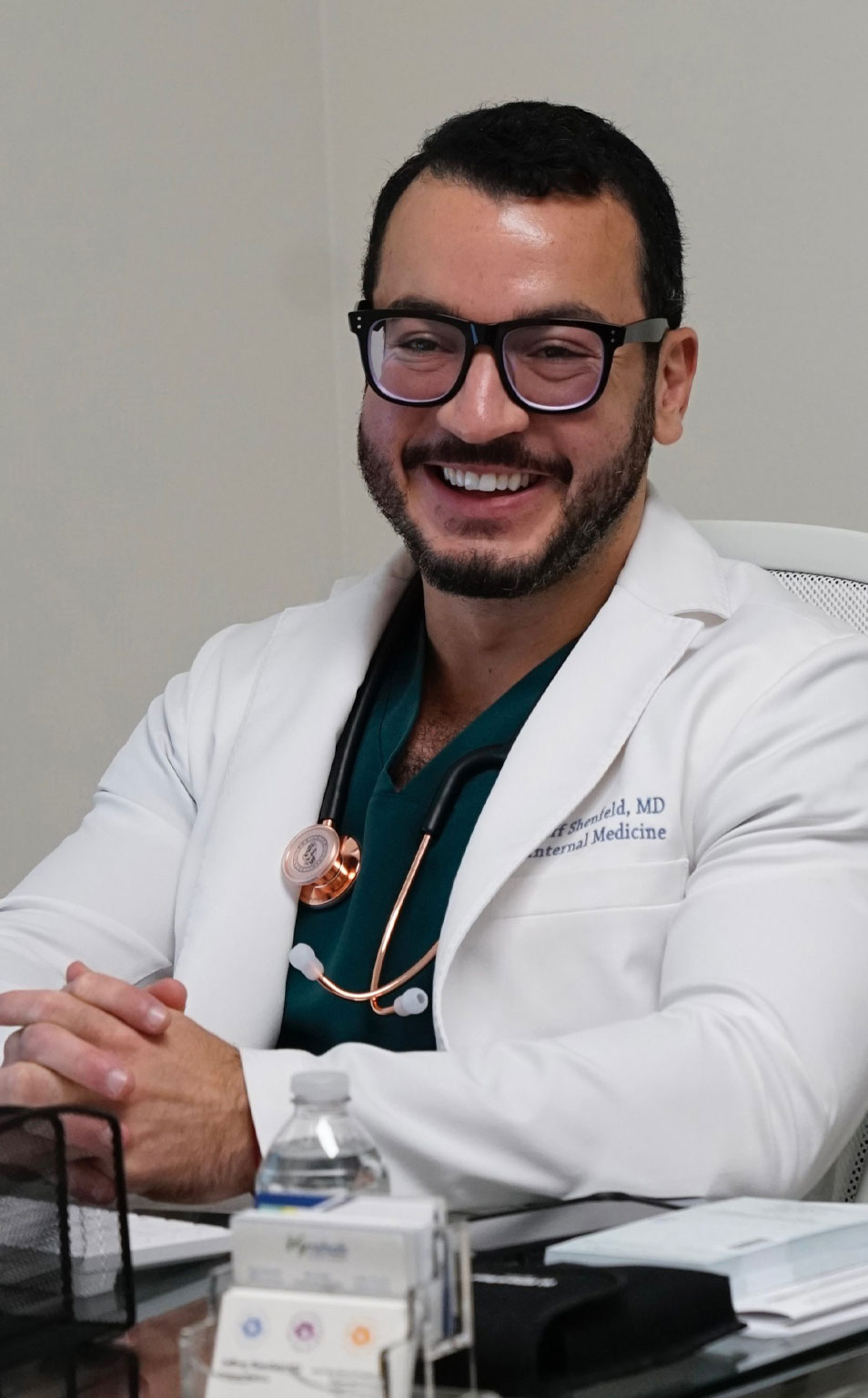 A man wearing glasses, smiling, sitting at a desk with a stethoscope around his neck.