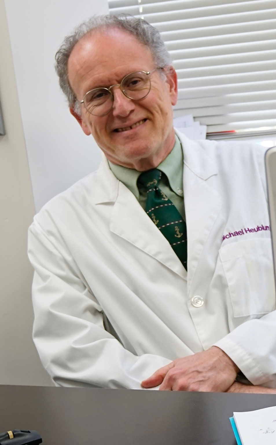 The image shows a man wearing a white lab coat and glasses, sitting at a desk with his arms crossed.