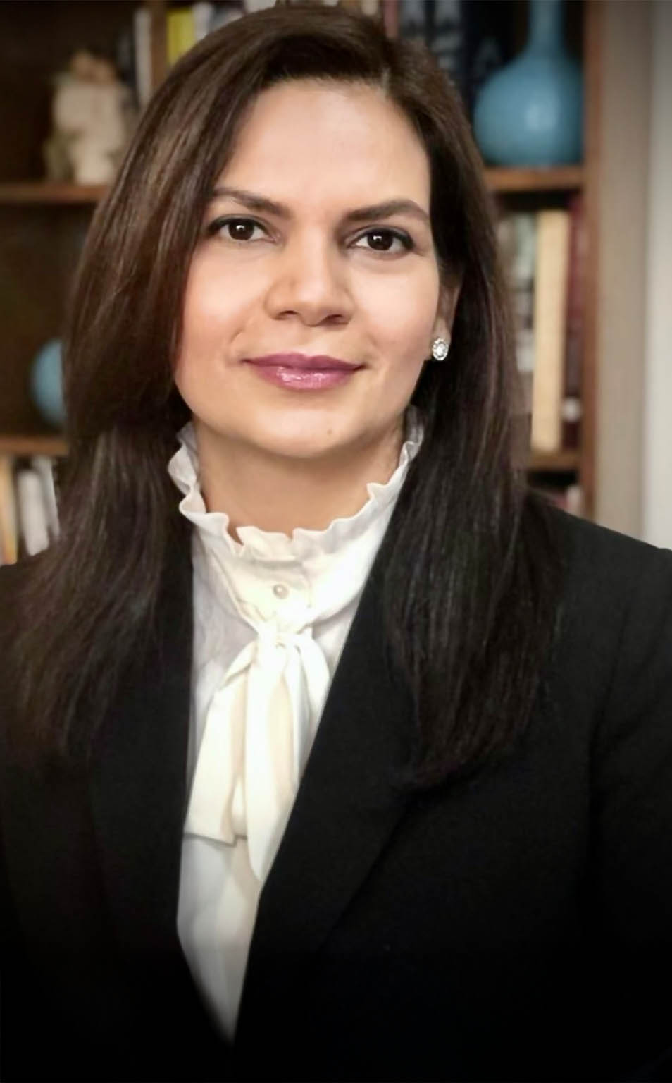 The image shows a woman posing for a portrait photograph with a professional appearance, wearing a suit and tie, against a blurred background that suggests an indoor setting with bookshelves and a vase.