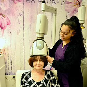 A woman with a head full of hair sits in a chair while another woman, standing behind her, uses a hair straightening device on her hair.